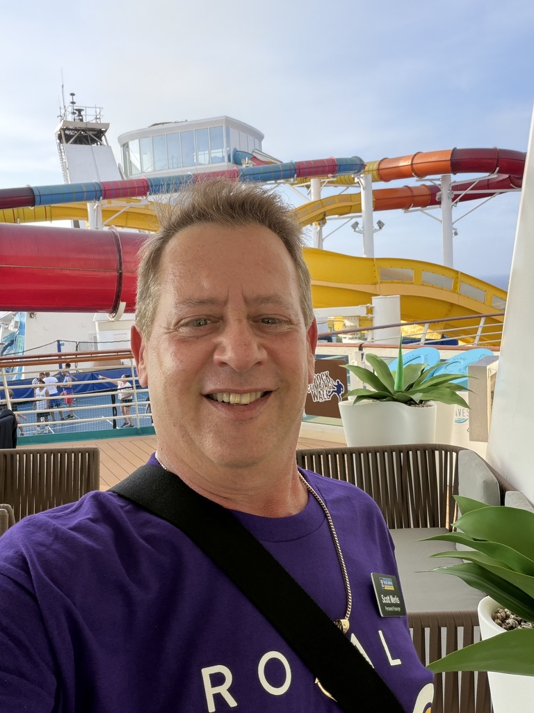 A bright day on the open deck: Scott A. Astin smiling alone on an open ship deck beneath the waterslides