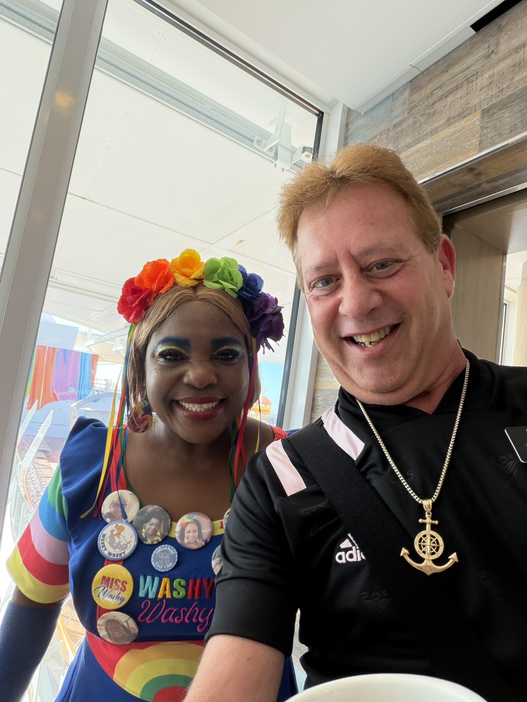 Color, joy, and a shipboard hello: Scott A. Astin smiling with a colorful shipboard performer wearing a flower headpiece and keepsake pins
